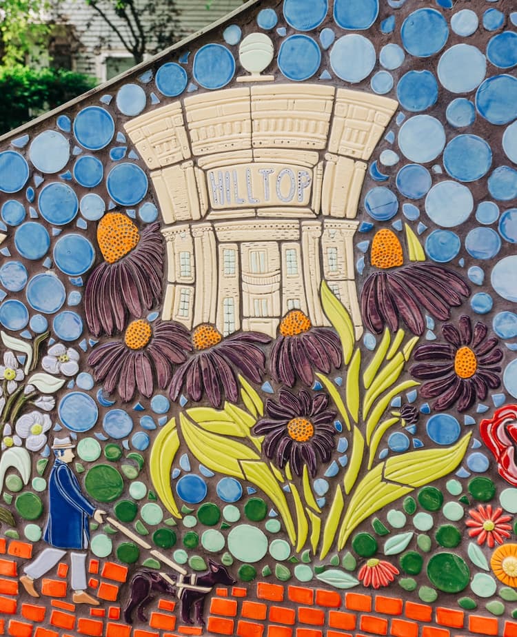 A mosaic panel of the creamy-white Hilltop Theatre building growing out of purple coneflowers, with a person walking a guide dog along the brick path below